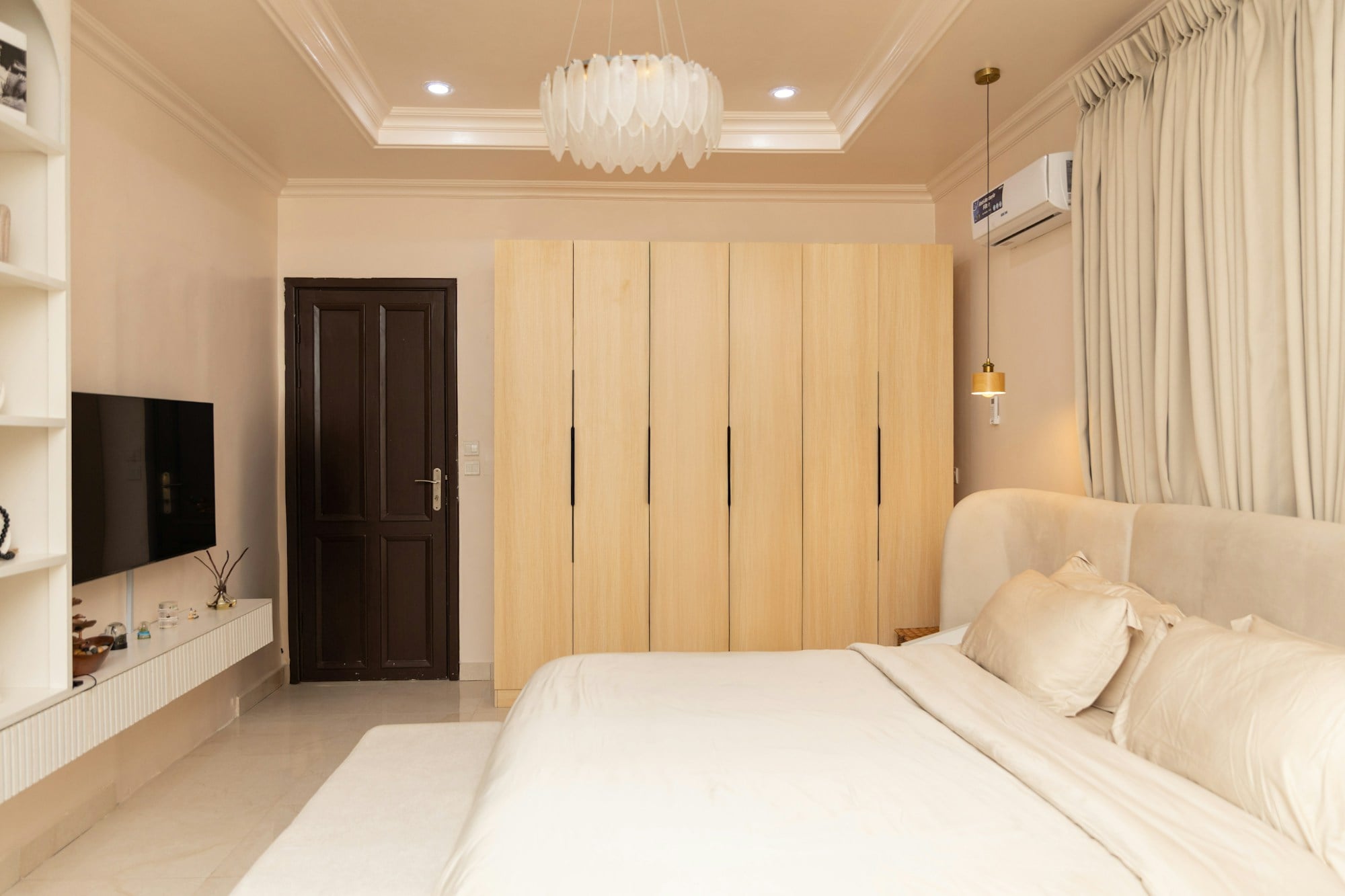 Bedroom with a run of pale oak wardrobe doors beside a cream upholstered bed, a pendant light and floor-length curtains