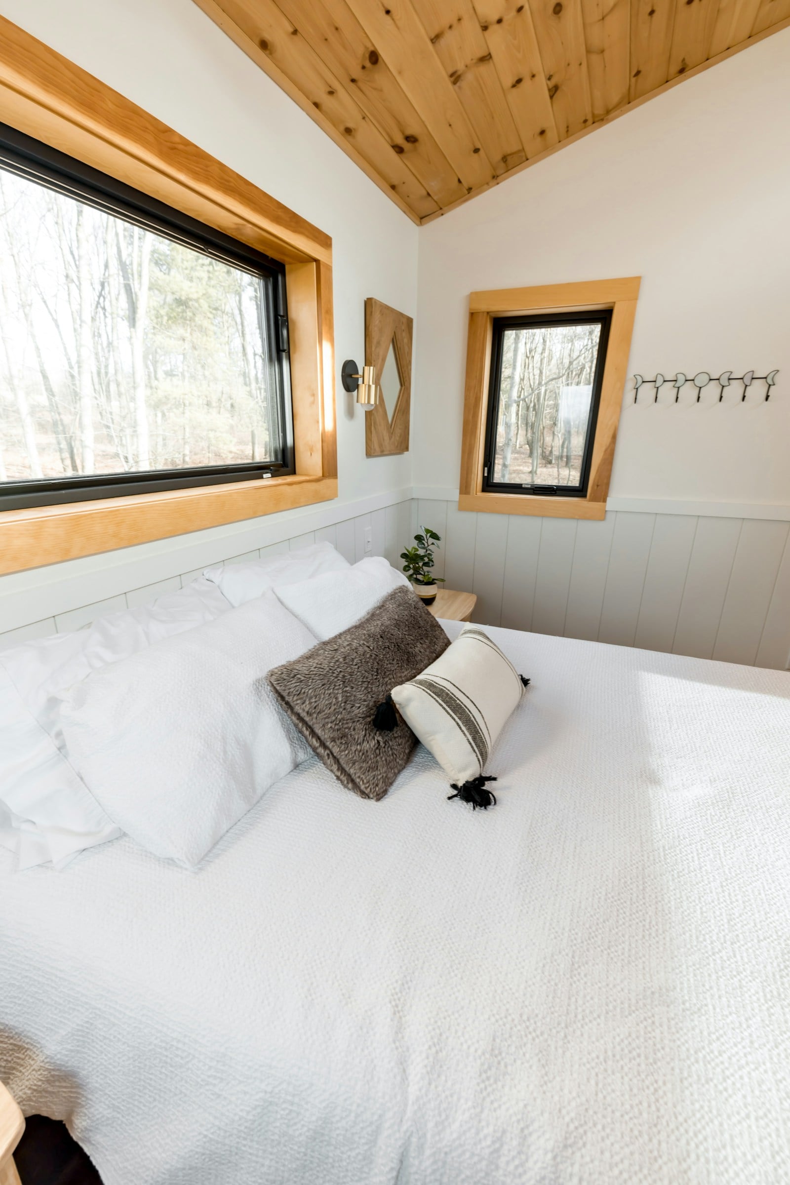 Bright bedroom with white bedding, a pine-lined sloping ceiling, timber-framed windows onto woodland and pale grey panelling
