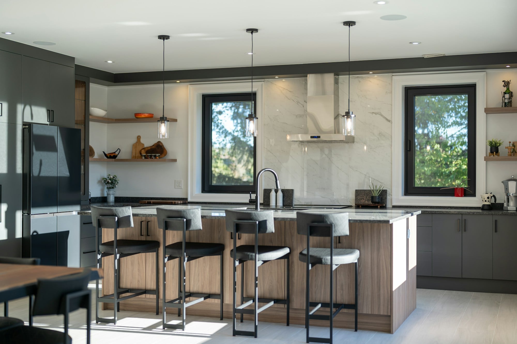 Bright kitchen with a timber-fronted island, four black bar stools, grey base cabinets, pendant lights and a marble splashback