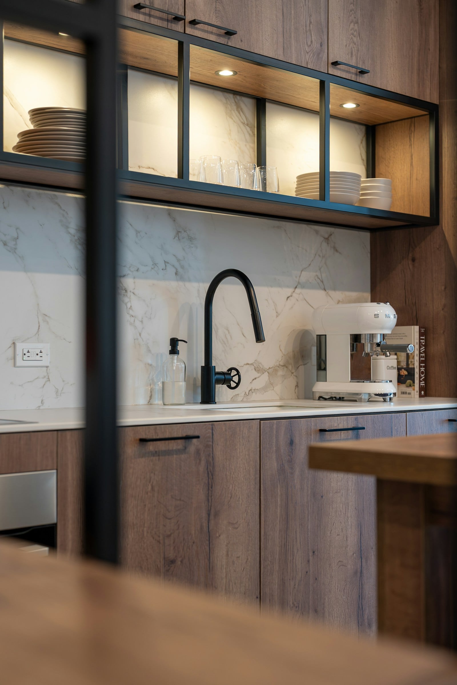 Close view of a kitchen with walnut-toned cabinetry, glazed wall units with built-in lighting, a marble splashback and a black pull-out tap