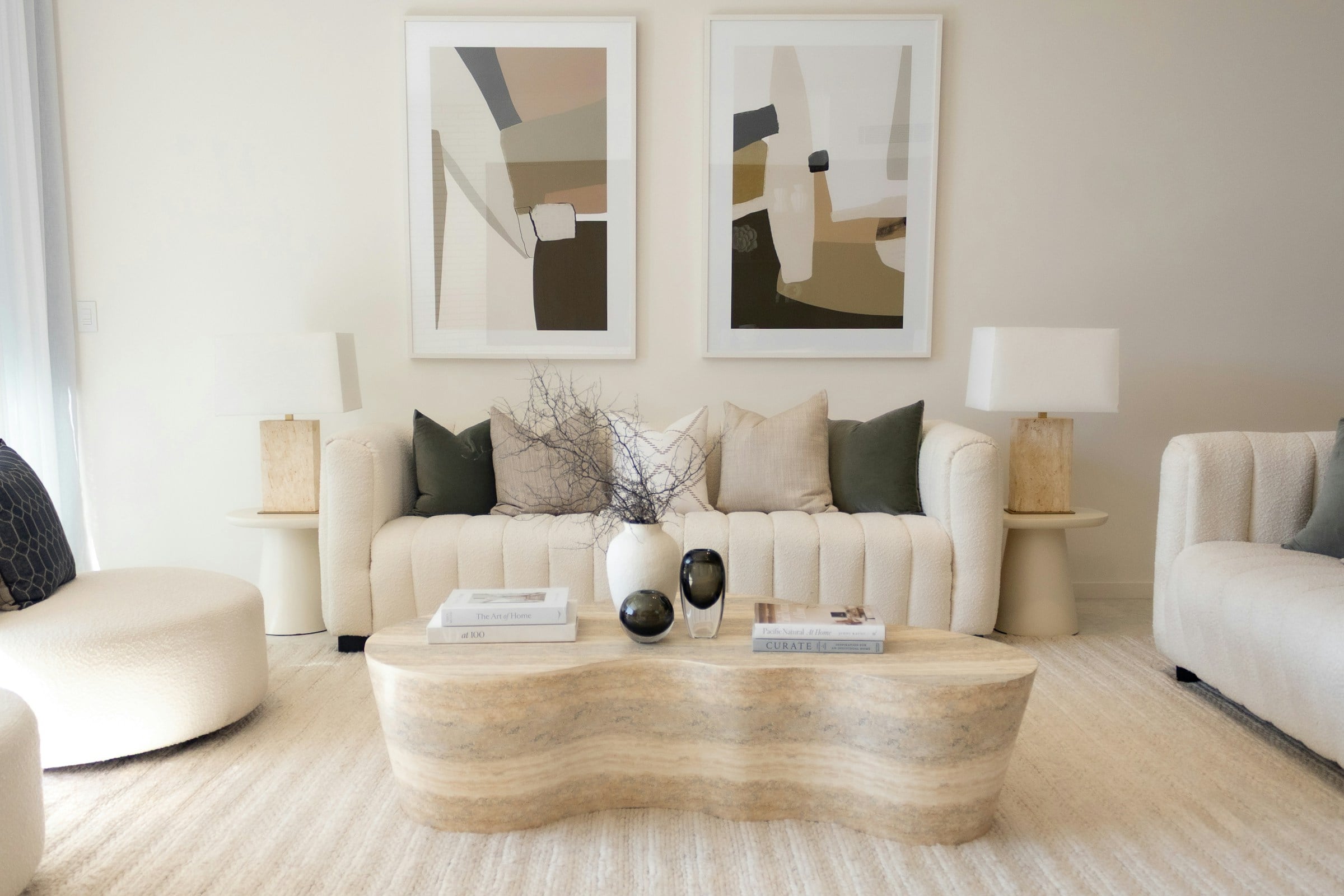 Calm living room with a cream channel-tufted sofa, a sculpted travertine coffee table, two abstract prints and soft neutral cushions