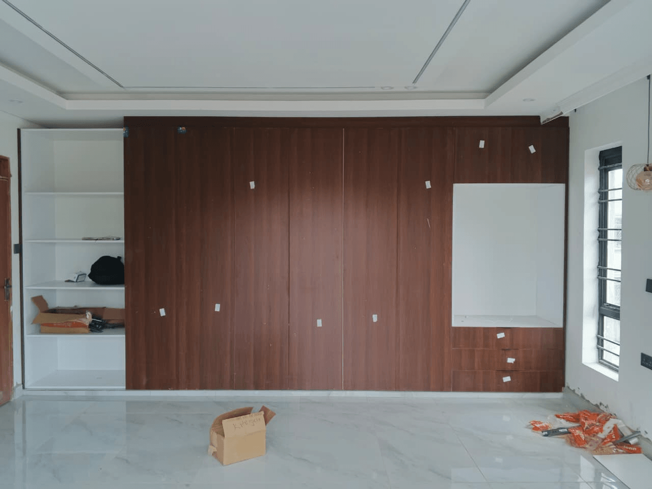 Full-width fitted storage wall with wood-tone doors, white open shelving on the left and a white recessed niche above three drawers on the right, in a room that is still being finished