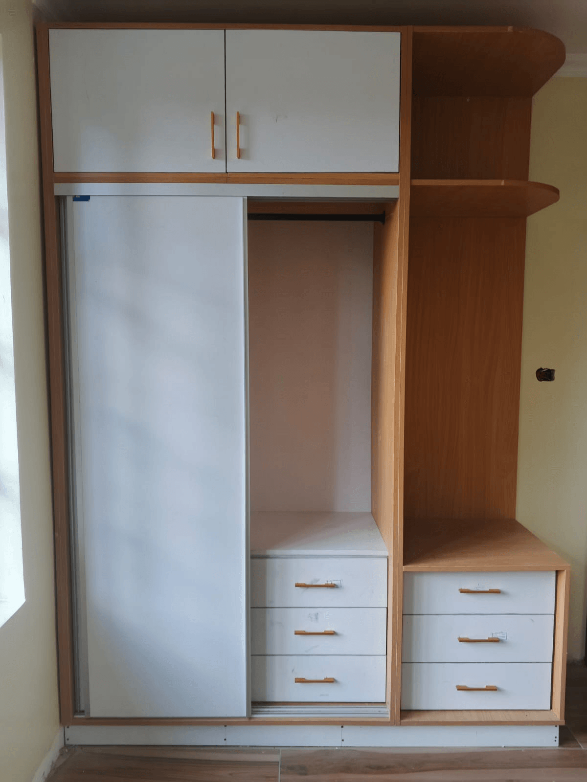 Fitted wardrobe with a white sliding door, a hanging rail, two banks of drawers with bar handles, overhead cabinets and open corner shelves in a light wood finish