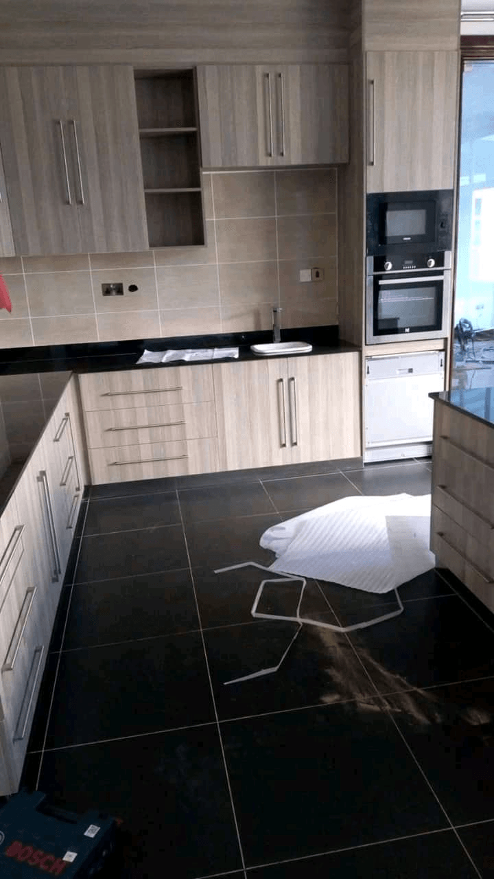 Fitted kitchen with light wood-grain cabinets, a black worktop, and a built-in oven and microwave above a dark tiled floor, still being finished